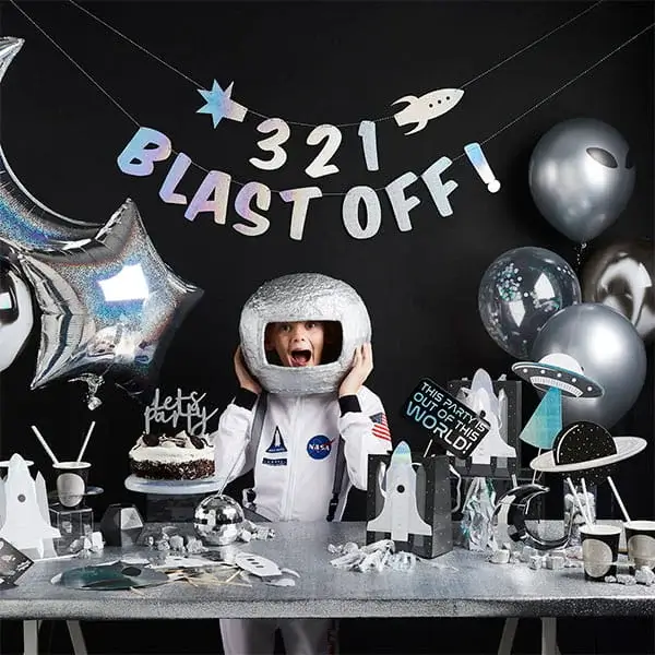 Outer space themed party setup with astronaut costume, rocket decorations, silver balloons, and a blast off banner on a dark background